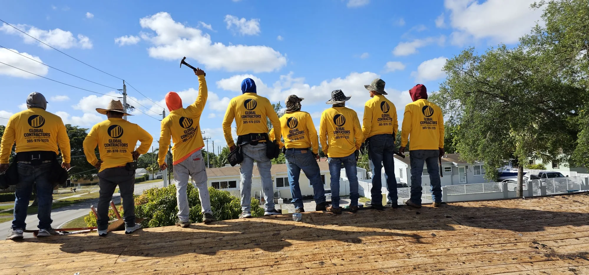 Ocho miembros de la cuadrilla de Global Contractors con camisas amarillas de la empresa en un techo, uno levantando un martillo